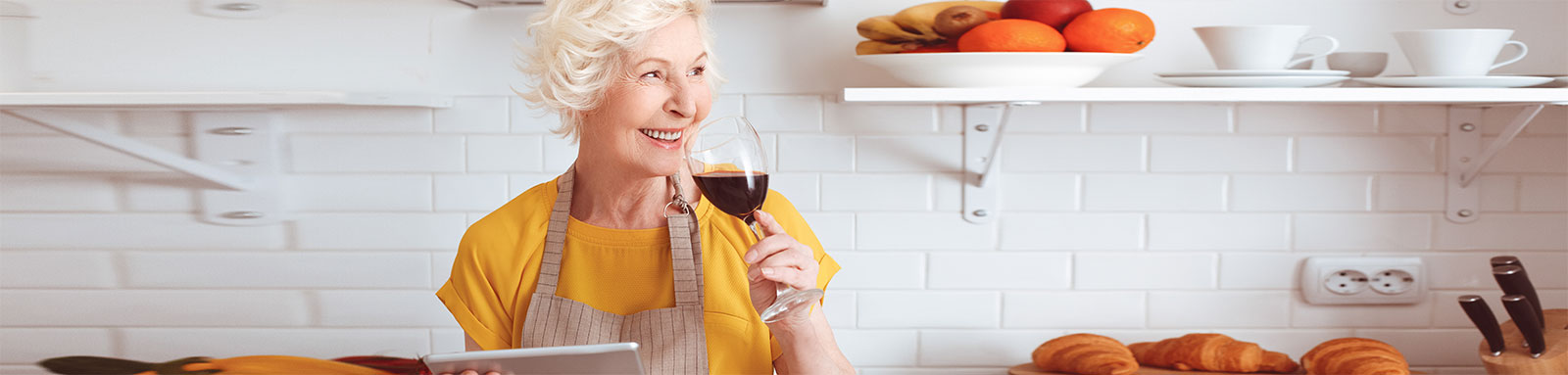 Elderly woman drinking red wine in the kitchen while holding tablet